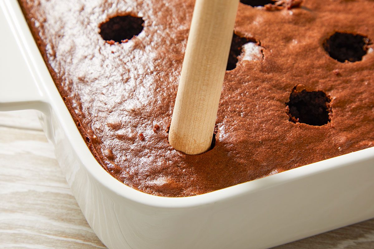 Overhead view of a baked chocolate cake with evenly spaced holes being poked using a wooden handle, preparing the cake to absorb the Irish cream filling.