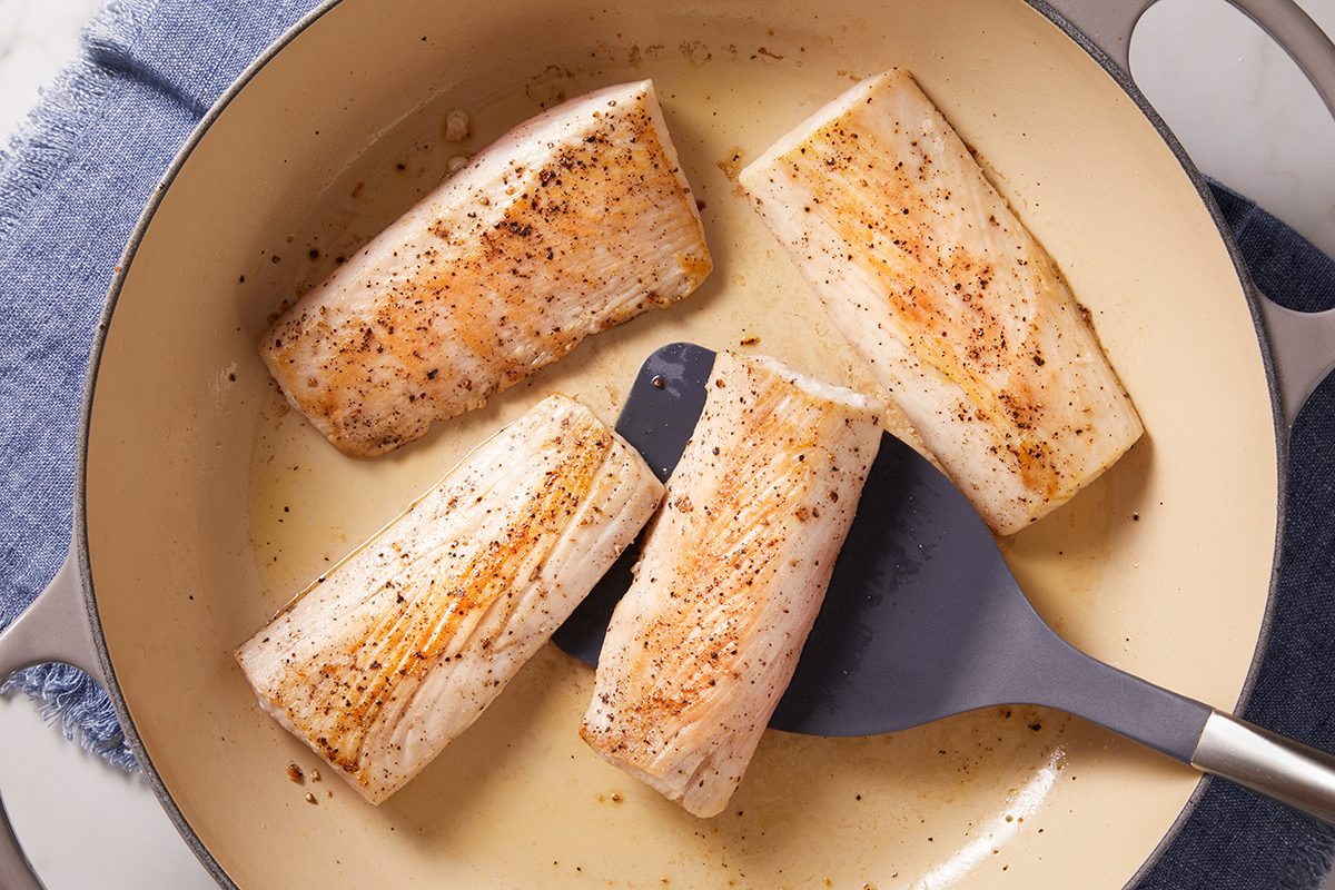 Three pieces of seasoned fish fillets are being cooked in a beige skillet. A spatula is lifting one piece, and the pan sits on a blue cloth.