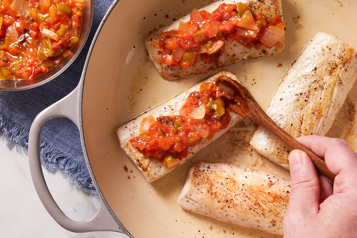 Four fillets of white fish are cooked in a pan, with a hand spooning chunky tomato and vegetable sauce over them. A bowl with extra sauce sits nearby on a blue cloth.