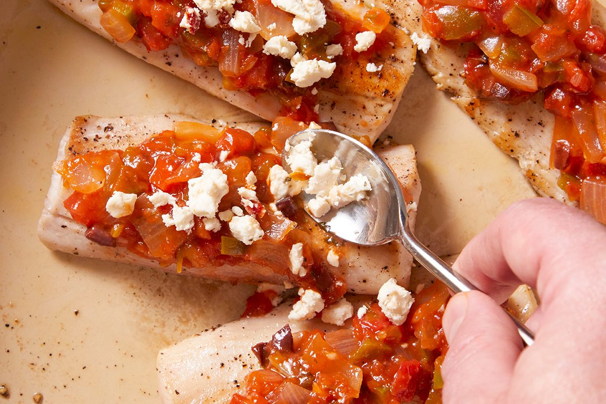 A hand uses a spoon to add crumbled cheese onto fish fillets topped with a chunky tomato and onion sauce in a pan.