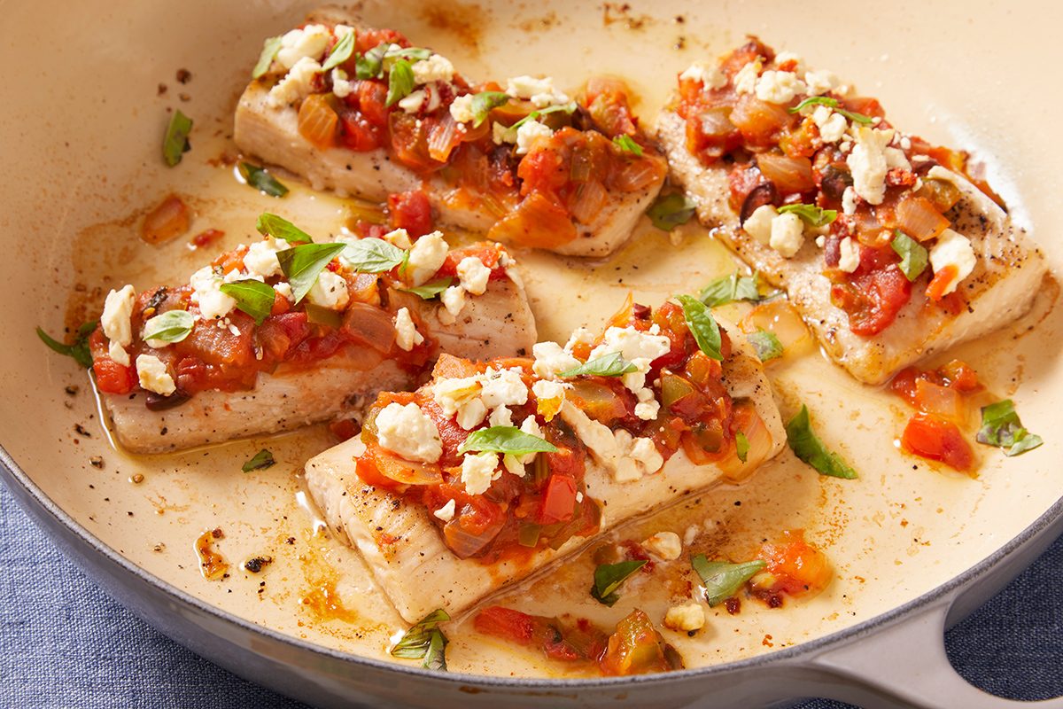 Four pieces of baked white fish topped with a tomato, herb, and feta cheese mixture, garnished with fresh herbs, served in a light sauce inside a baking dish.