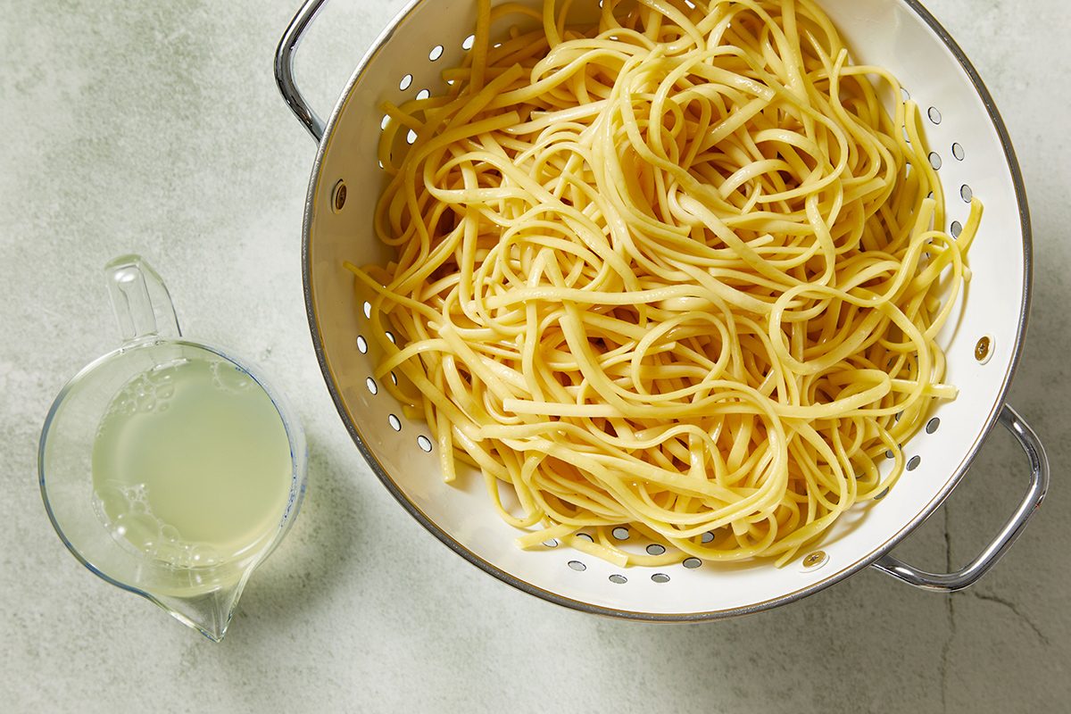 A colander filled with cooked linguine pasta sits on a light surface next to a glass measuring cup containing pasta water.