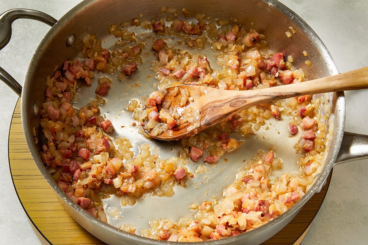 Chopped onions and diced pancetta are being sautéed in a stainless steel pan. A wooden spoon is stirring the mixture, which is lightly browned and sizzling. The pan rests on a wooden trivet.