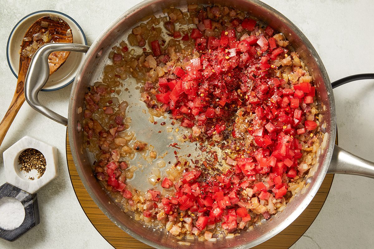 A saucepan with diced tomatoes, onions, and spices being sautéed, next to a wooden spoon, a small dish with red flakes, and a container of salt and pepper on a white surface.