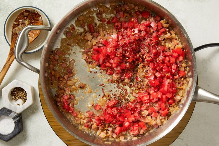A saucepan with diced tomatoes, onions, and spices being sautéed, next to a wooden spoon, a small dish with red flakes, and a container of salt and pepper on a white surface.