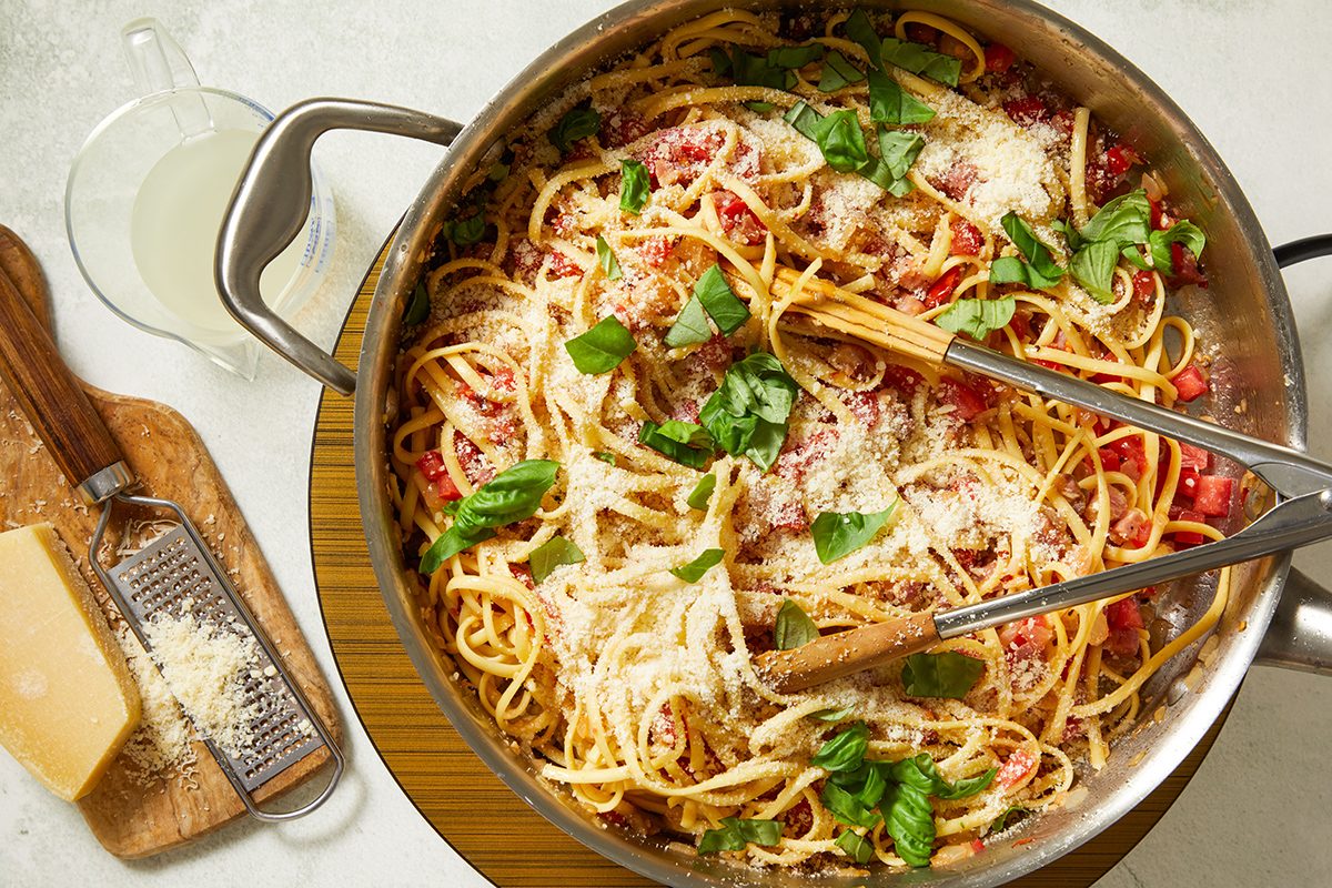 A pot of spaghetti topped with grated cheese and fresh basil, with wooden tongs resting inside. Nearby, a block of cheese and grater sit on a cutting board, and a glass of water is visible in the background.