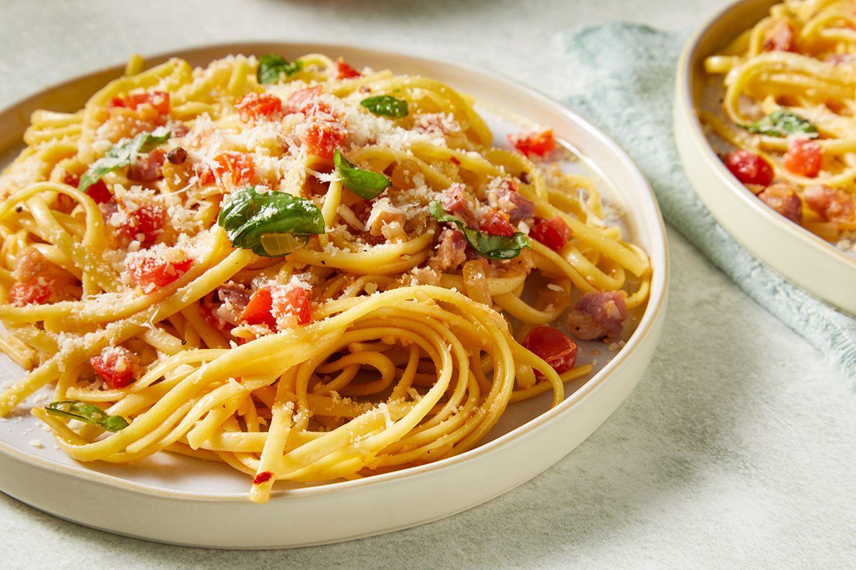 A plate of spaghetti topped with grated cheese, diced tomatoes, fresh basil leaves, and bits of bacon or ham, served on a light-colored dish with another plate of pasta in the background.