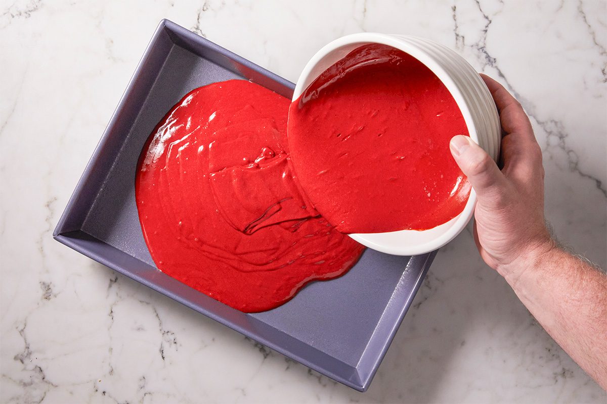 A overhead, horizontal shot of Red Velvet Cake bites red frosting being added to a rectangular pan as it is poured from a white bowl from a hand entering the image on the right.
