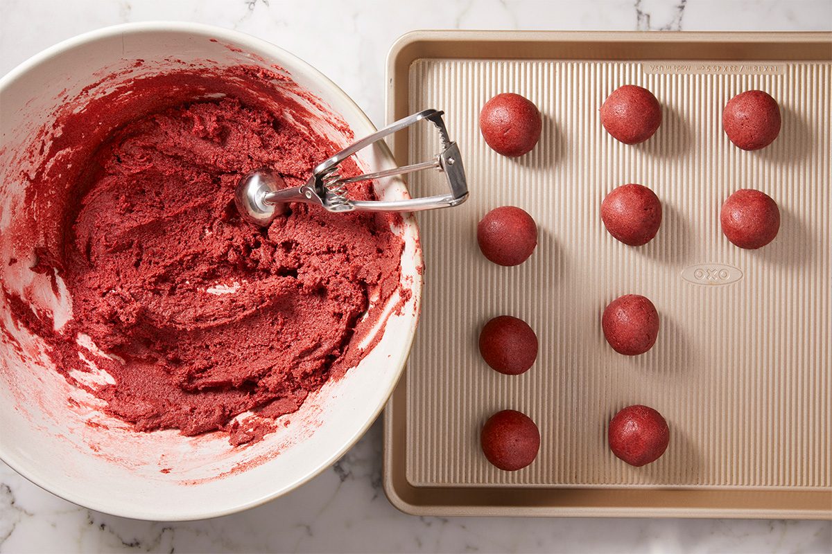 A overhead, horizontal shot of Red Velvet Cake bites being prepped from a bowl of cake batter, scooped with a round scoop—cake bite balls are then placed on the baking sheet