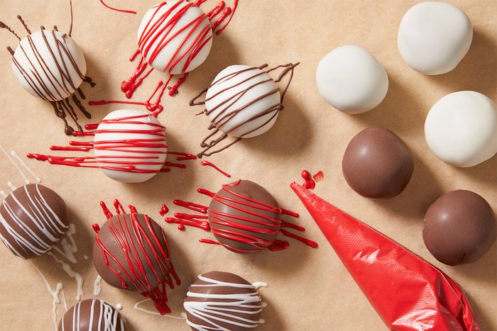 A overhead, horizontal shot of Red Velvet Cake bites on parchement paper being decorated with red frosting in a tube—some undecorated Cake Bites remain in the upper right of the image.