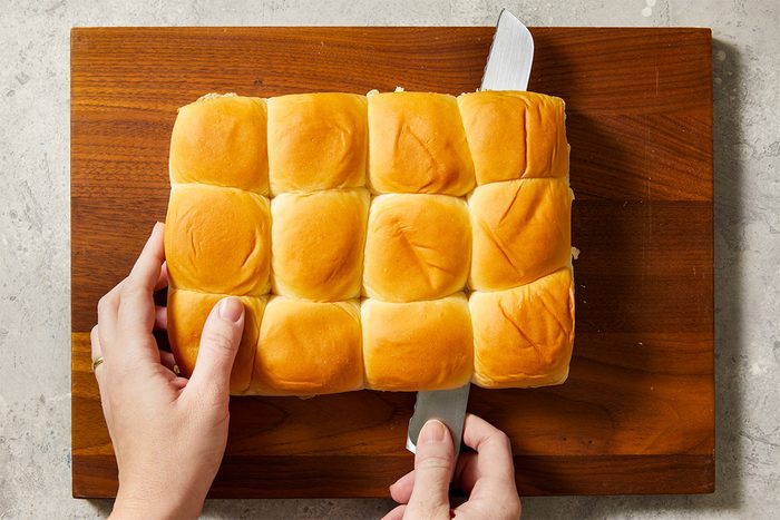 Overhead step-by-step food photography showing a slab of Hawaiian sweet rolls being sliced horizontally with a knife to prepare Reuben sliders.