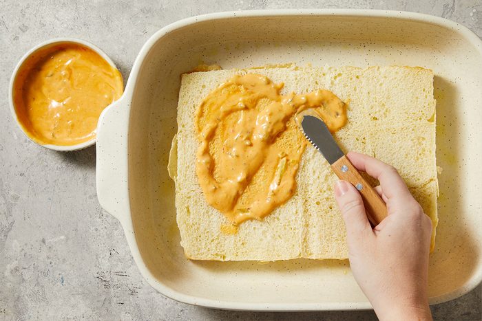 Overhead step-by-step food photography showing Russian dressing being spread evenly over the bottom half of sliced Hawaiian rolls in a baking dish.