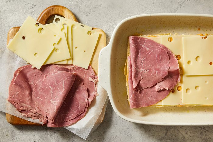 Overhead step-by-step food photography showing sliced Swiss cheese and thinly sliced corned beef layered over rolls for Reuben sliders.
