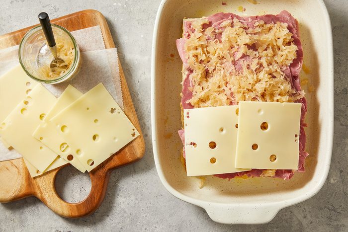Overhead step-by-step food photography showing sauerkraut and Swiss cheese layered over corned beef on slider rolls before topping with bread.