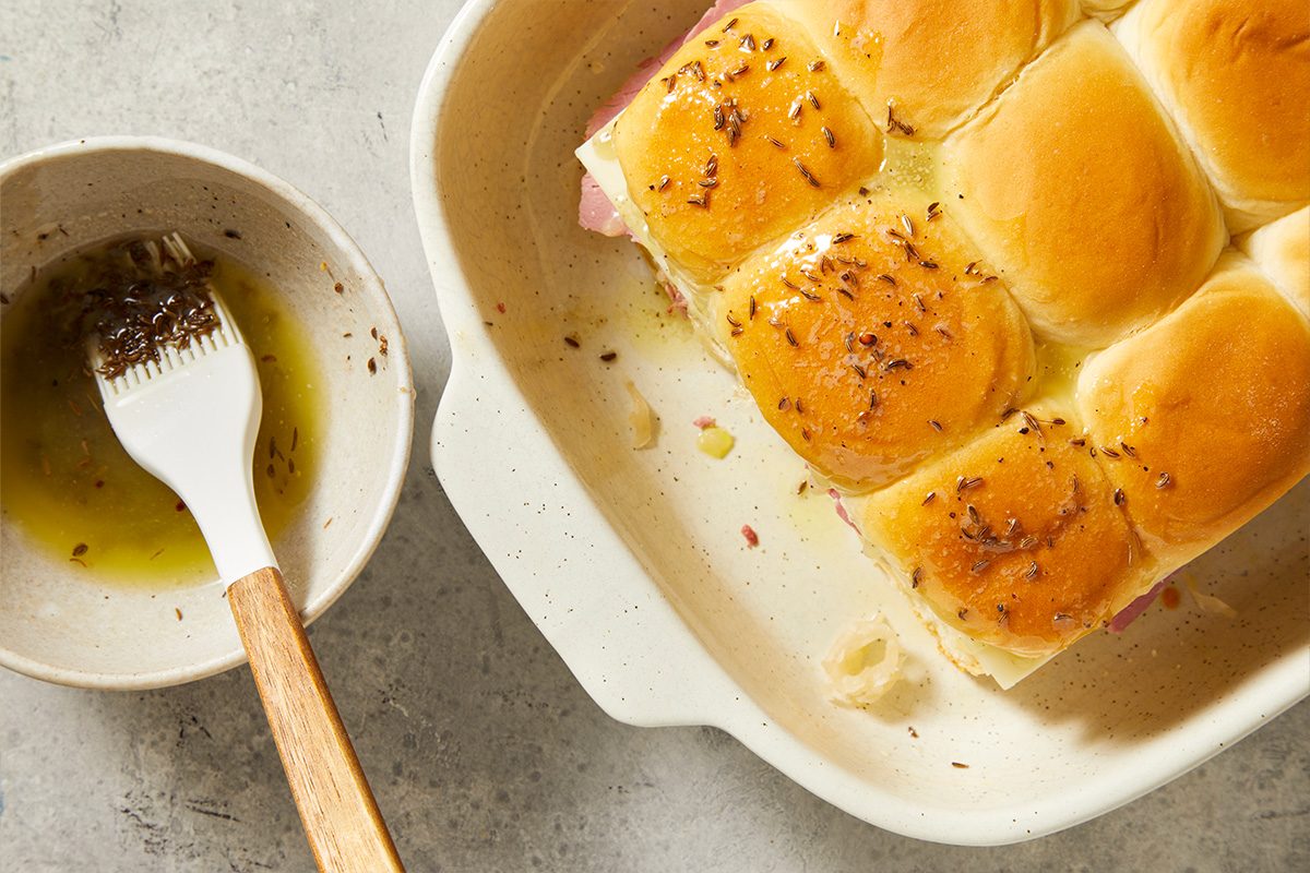 Overhead step-by-step food photography showing melted butter being brushed over assembled slider tops and sprinkled with seasoning.