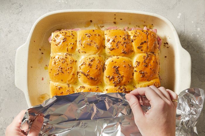 Overhead step-by-step food photography showing assembled Reuben sliders being covered with foil in a baking dish before baking.