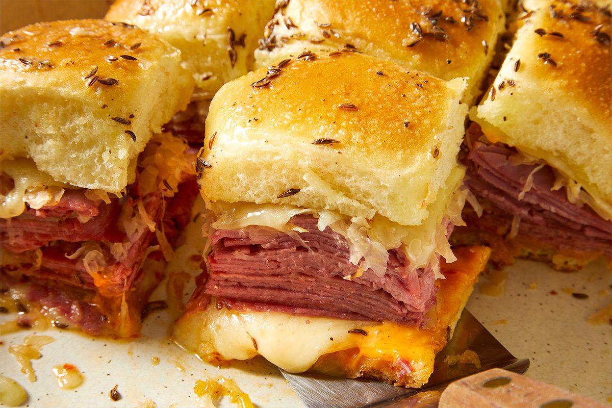 Overhead close-up food photography showing Reuben sliders being cut, revealing layers of corned beef, Swiss cheese, and sauerkraut.