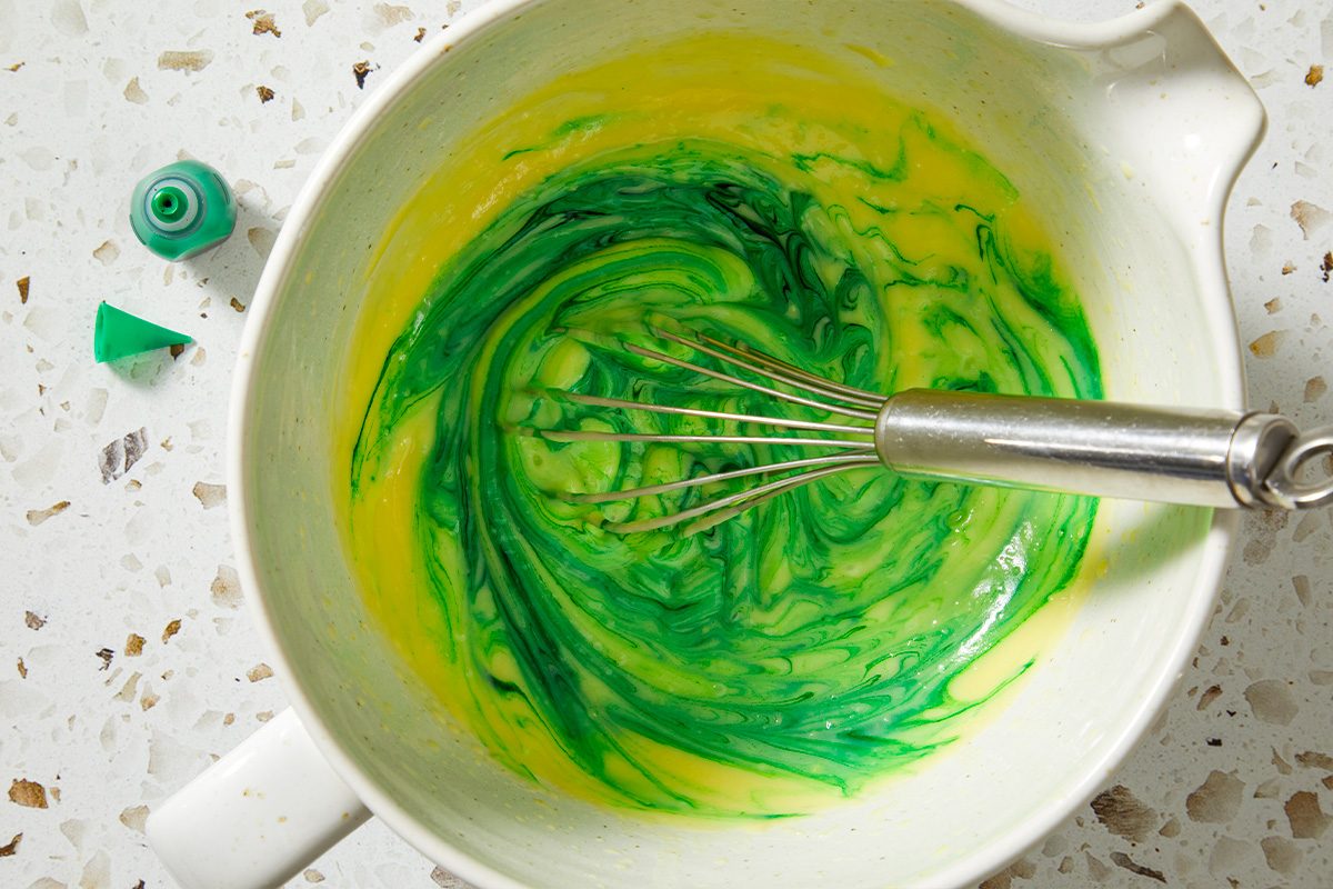 Overhead, horizontal image of a bowl as green food coloring is whisked into the mixture. The vivid green color begins to develop, signaling the signature Shamrock Shake appearance during this step-by-step process.