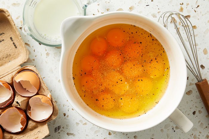 Overhead shot of a mixing bowl filled with cracked eggs, surrounded by broken eggshells and a whisk, ready to be poured into the slow cooker mixture. {