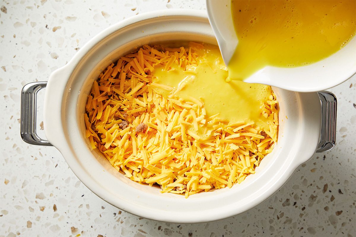 Overhead shot of beaten eggs being poured into the slow cooker over a generous layer of shredded cheese, creating the base for the breakfast burrito filling.