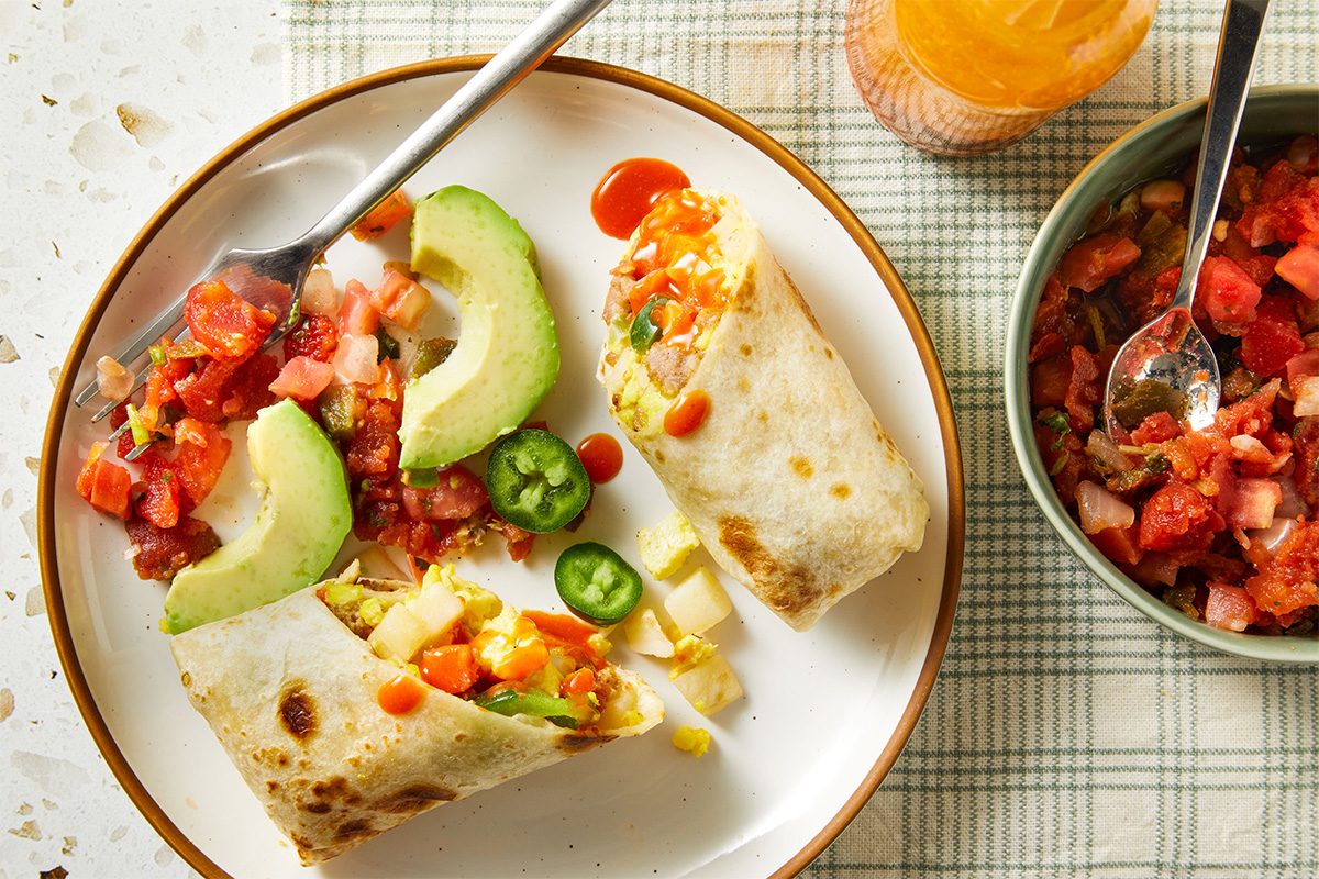 Overhead shot of sliced Slow-Cooker Breakfast Burritos on a white plate, served with salsa, avocado wedges, and jalapeños, creating a colorful finished dish.