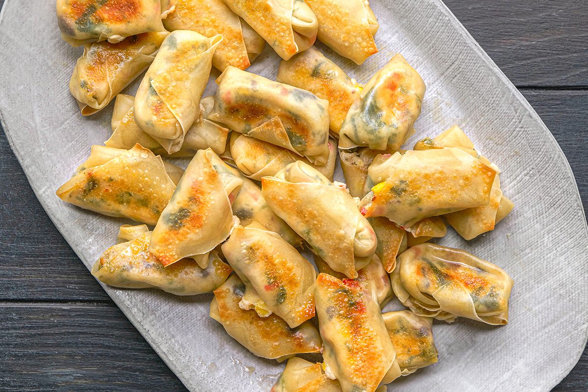 A platter filled with neatly arranged, golden-brown baked pastry rolls, likely filled with a savory mixture, on a light gray textured plate set atop a dark wooden surface.