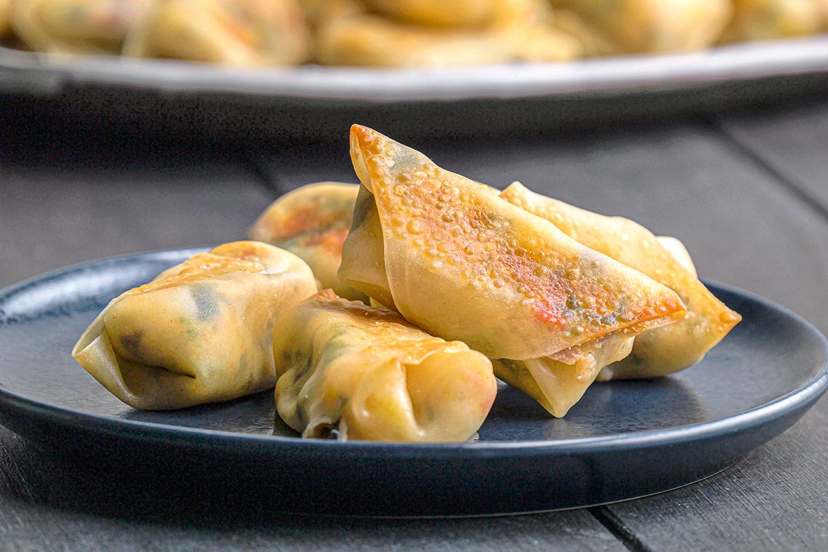 Three crispy, golden-brown spring rolls are arranged on a dark plate, with more spring rolls blurred in the background. The filling is slightly visible through the thin wrappers.