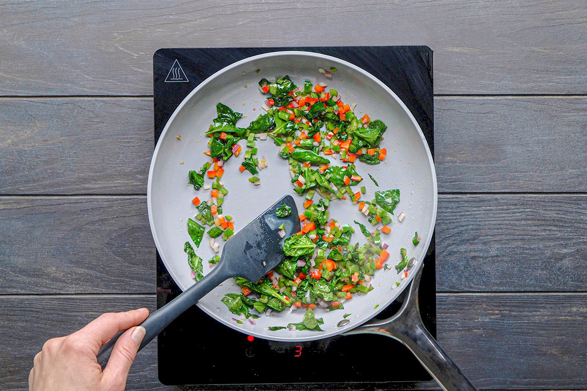 A hand stirs chopped green spinach and diced orange carrots in a white frying pan on a stovetop.