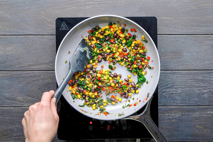 A hand stirs a mixture of black beans, corn, spinach, and diced red peppers in a skillet on an electric stovetop.