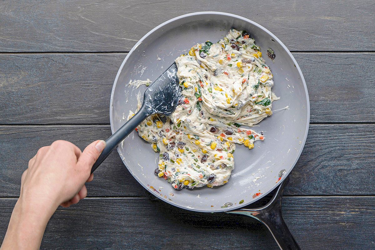 A hand uses a spatula to stir a thick mixture with colorful bits, possibly vegetables, in a frying pan on a dark wooden surface.