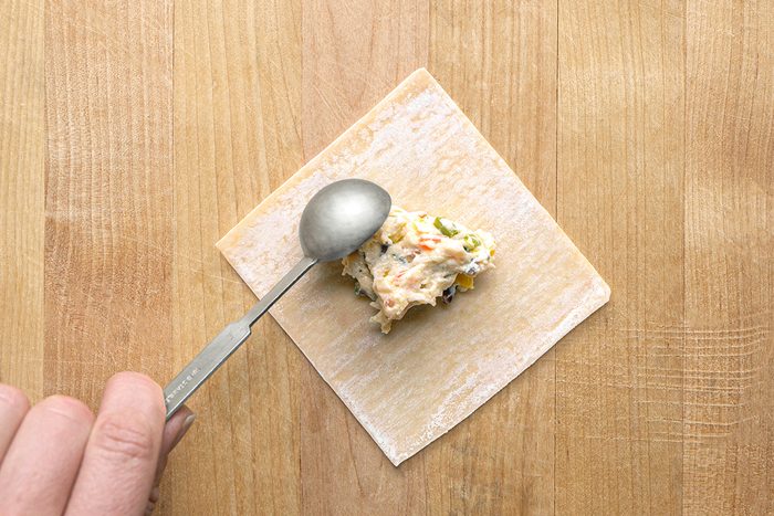 A hand holding a spoon places a dollop of creamy filling onto the center of a square wonton wrapper on a wooden surface.