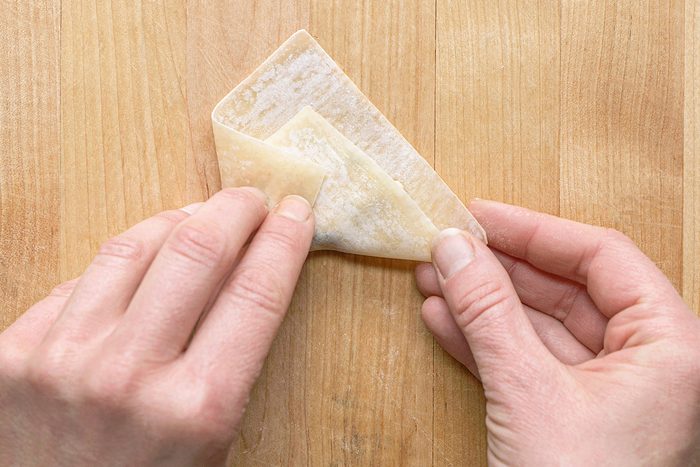 Two hands folding a wonton wrapper into a triangle shape on a wooden surface.