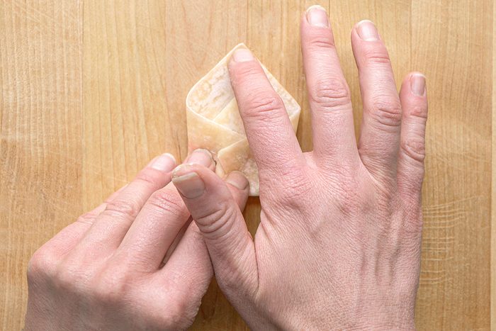 Two hands folding a wonton wrapper on a wooden surface, shaping it into a triangle.