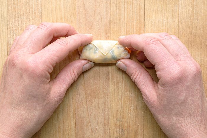Two hands folding a wonton or dumpling wrapper on a wooden surface, with the wrapper partially folded and the filling visible inside.