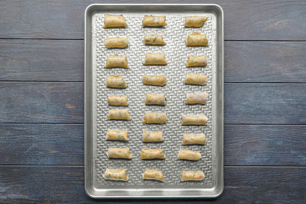 A baking sheet with neatly arranged, evenly spaced, uncooked stuffed pasta pieces on a textured metal tray, placed on a dark wooden surface.