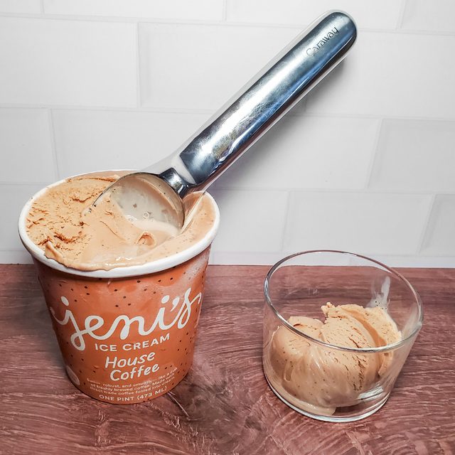 A container of Jeni&rsquo;s House Coffee ice cream with a scoop inside is next to a small glass bowl holding a scoop of the ice cream, all on a wooden surface with a white tiled background.