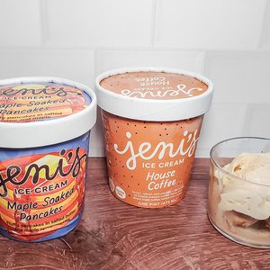 Two pints of Jeni’s ice cream, flavors Magic Stoopwafels Pancakes and House Coffee, sit on a counter next to a glass bowl containing scoops of ice cream. A white tiled wall is in the background.