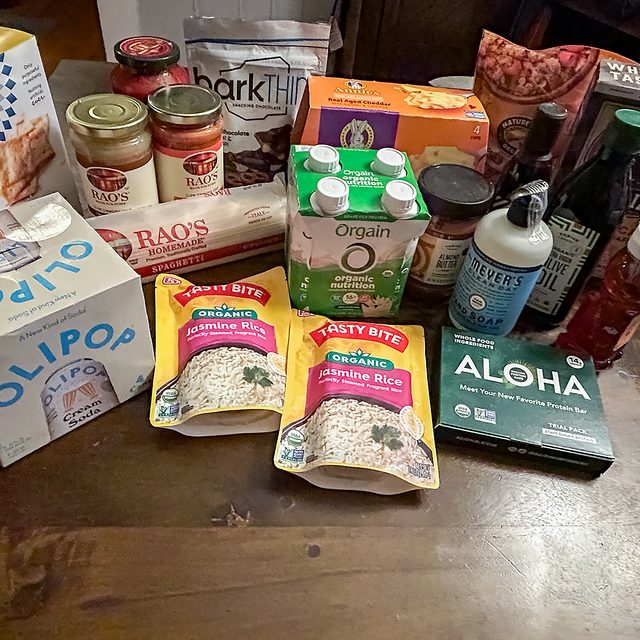 A variety of groceries on a table, including Rao&rsquo;s pasta sauce, barkTHINS chocolate, Olipop soda, Tasty Bite rice packs, Orgain protein shakes, Aloha protein bars, and several canned and bottled items.