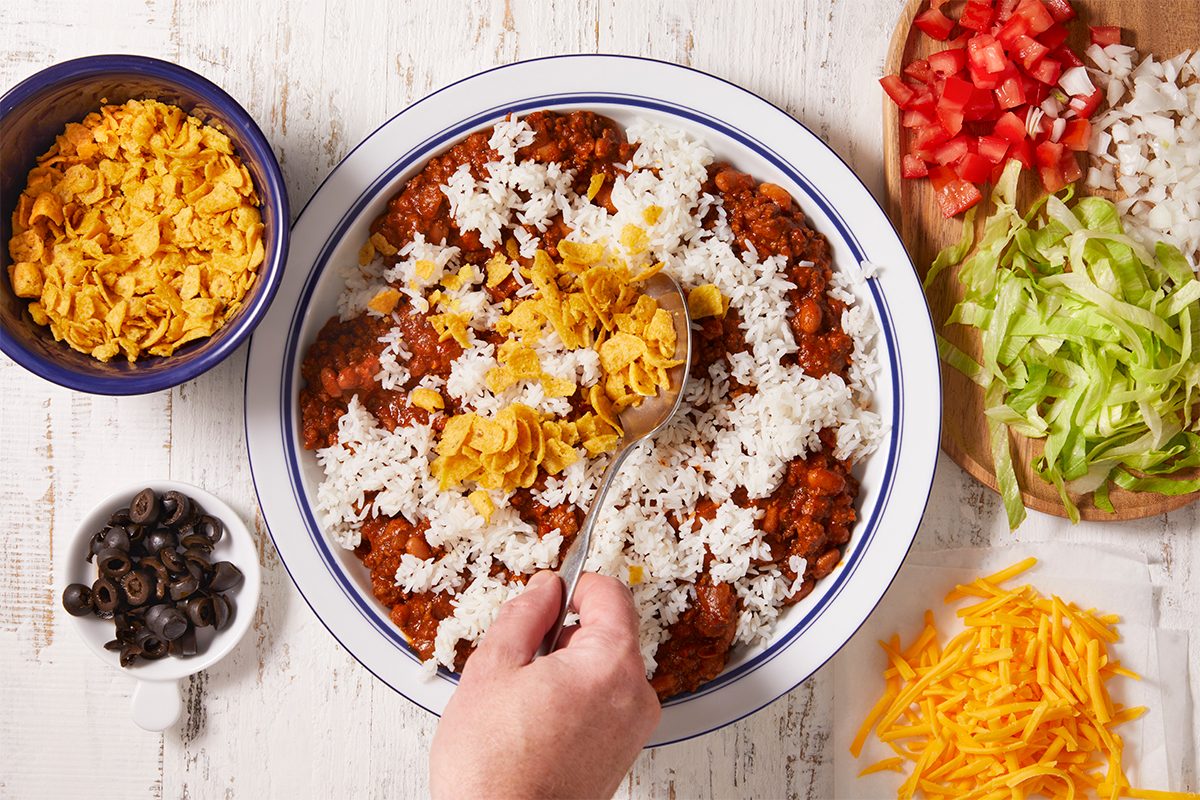 On a platter, layer the meat mixture, rice, corn chips, cheese, onion, lettuce, tomatoes and olives.