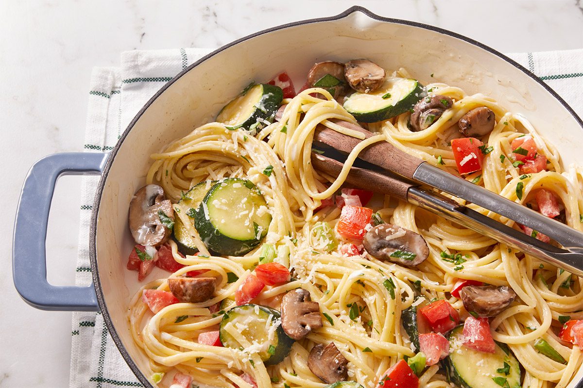 Overhead shot of a pan filled with Vegetable Linguine mixed with sautéed zucchini, mushrooms, and tomatoes, with stainless steel tongs placed across the pasta.